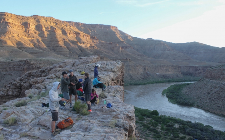 A group of people stand on a cliff, overlooking a river winding through a desert landscape.